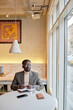 © Seventyfour - Man wearing sunglasses and beige turtleneck sweater writing notes while sitting at a table by large window in cafe. Modern interior with artistic decor on wall present