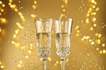 Glasses of sparkling wine and confetti against golden background with blurred lights, closeup. Christmas decor