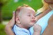 © malyutinaanna - A peaceful baby nestles in a mother’s embrace, enjoying a tranquil nap beneath a clear blue sky, surrounded by vibrant greenery and gentle sunshine.