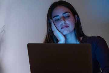 Late-night portrait of a tired and exhausted woman working on her laptop, showing boredom and frustration. Alone at home, she struggles with insomnia while staring at the computer, unable to sleep 