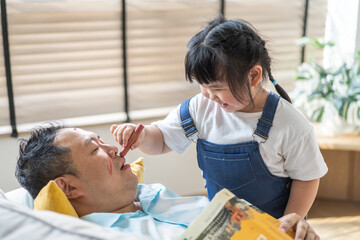 Asian father asleep on couch while little daughter playfully applies makeup on his face, funny family moment playful asian child, humor, bonding, daddy and daughter love, parenting, relaxation at home