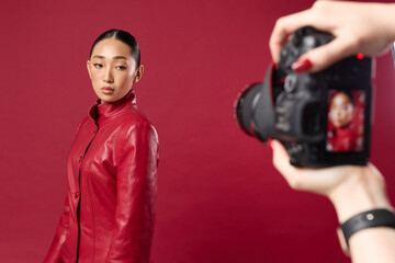 Wall Mural - Fashionable asian brunette woman in red leather jacket taking a photo with digital camera on red background