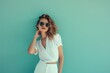 © Maria - Beautiful young girl with model appearance in sunglasses and white dress posing on turquoise background