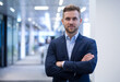 © Arshadul - Portrait of a confident young male entrepreneur in a suit, standing in the office with his arms crossed, smiling directly at the camera.