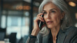 © Nattapol - An older businesswoman with silver hair is engaged in phone conversation, showcasing focused and professional demeanor in modern office setting