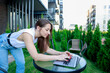 © Aleksandra - young woman casual attire works on laptop outdoors leaning over glass table She’s in a modern urban garden setting combining work leisure. Ideal for remote work, freelancer, or digital nomad themes.