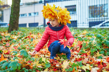 Adorable preschooler girl in maple leaves head decoration enjoying nice and sunny autumn day outdoors. Happy child walking on a fall day in Paris, France.