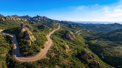 Sticker - Aerial view of a winding road through a mountainous landscape with rocky formations, dense green vegetation, and clear blue skies.