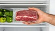 © lililia - A hand grips a frozen meat package beside an open refrigerator showcasing a variety of fresh produce and meats, ready for delicious meals and cooking adventures