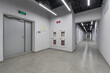 © alhim - A corridor with white brick walls in an office building. A gray door with a green emergency exit sign.