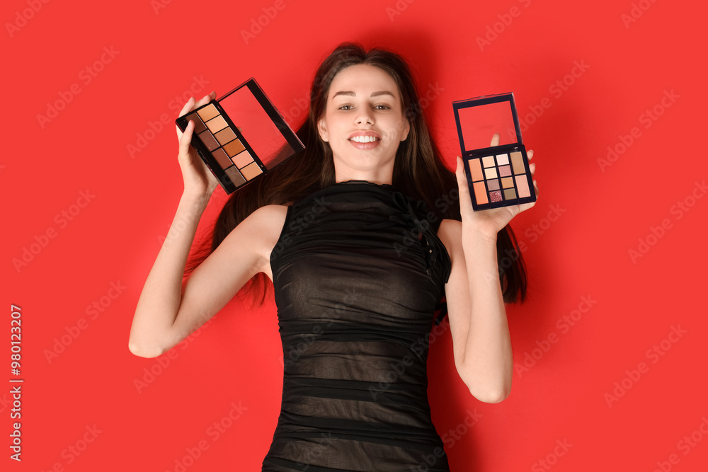 Happy smiling young woman with eyeshadows palettes lying on red background