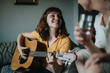 © qunica.com - Smiling woman in a yellow shirt plays guitar while sitting on a sofa, spending quality time with a friend in a cozy home setting.