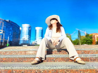 A woman sits comfortably on stone stairs in a city, enjoying the warmth of a sunny day while wearing a wide-brimmed hat