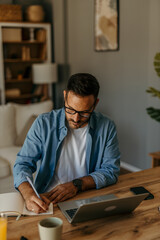 Handsome focused man in a denim shirt sitting at home and using a laptop