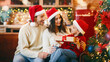 © Prostock-studio - Sweet girl in santa hat asking parents permission to open xmas gift, sitting next to christmas tree at home together