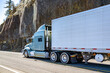 © vit - Powerful extended cab grey big rig semi truck tractor transporting cargo in refrigerator semi trailer running on the mountain highway road with rock cliff on the side