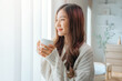 © oatawa - Attractive lady enjoying her morning coffee or tea at home, Smiling woman holding cup of tea in hands while looking out the window at morning.