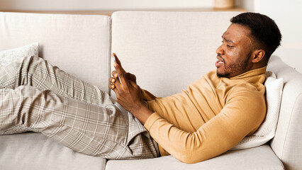 Canvas Print - Discontented Black Man Using Smartphone And Frowning Texting Or Browsing Internet Lying On Couch Indoor. Selective Focus