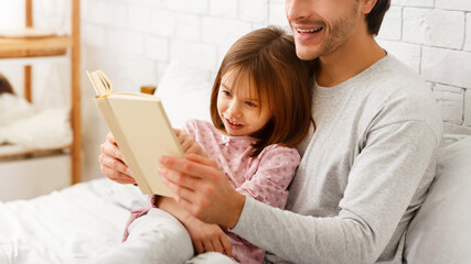 Wall Mural - Close up of preschool cute girl reading book with father, sitting on bed together