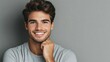 © fotofabrika - A young man smiles confidently while casually resting his hand on his chin against a gray background in a relaxed indoor setting