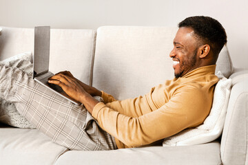 Wall Mural - Happy African American Man Using Laptop Computer Lying On Couch Indoor. Weekend At Home Concept.