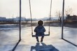© Janyawatt - Rear view of a child sitting on a swing in an empty playground