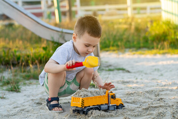 Little cute caucasian toddler boy of three and a half years old plays in the sandbox on a sunny summer day. Outdoor creative activities for kids. Toy dump truck and shovel.