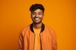 © Markus Schröder - Portrait of a smiling indian man in his 20s wearing a trendy bomber jacket isolated on soft orange background