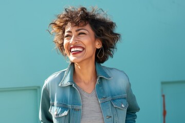 Wall Mural - Portrait of a cheerful indian woman in her 50s sporting a rugged denim jacket isolated in soft teal background