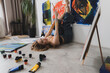 © Mihail - young woman lies on her back, legs raised, with her feet leaning against the wall, while painting in her studio. The space is filled with colorful art supplies and paintings