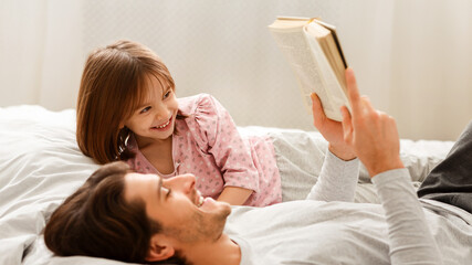 Wall Mural - Young man reading book to his little smiling daughter, laying on bed together