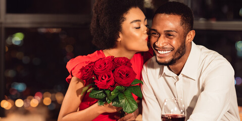 Happy Love Relationship. African American Girlfriend Kissing Boyfriend After Receiving Beautiful Bouquet Of Roses During Romantic Date In Restaurant.