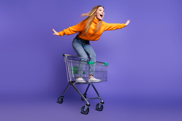 Poster - Photo of blonde hair cheerful youngster girl in orange turtleneck and jeans riding supermarket cart flying isolated on purple color background