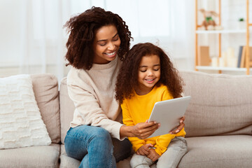 Wall Mural - Cute Black Family Of Mother And Daughter Using Tablet Computer Watching Cartoons Together Sitting On Couch At Home.