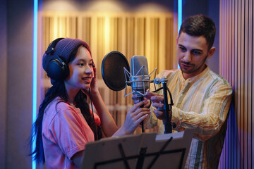 Two individuals in recording studio adjusting microphone setup with soundproof walls in background, wearing headphones and focusing on music production