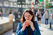© Connect Images - Woman in blue jacket eating a sandwich on a busy city street.