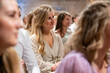 © Connect Images - Smiling young woman in a beige cardigan sitting among a crowd of people at an indoor event.