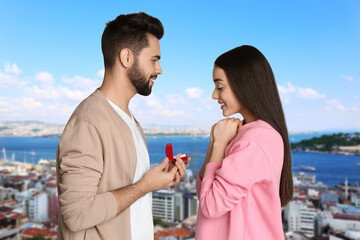 Poster - Man with engagement ring making marriage proposal to his girlfriend. Couple against cityscape