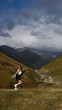© Vasya - A young woman leaps joyfully on a hillside, surrounded by vast mountains under a cloudy sky. A soft rainbow arcs in the background, adding a touch of magic to the dramatic scenery.
