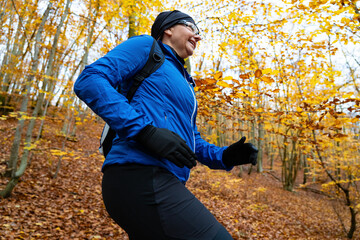 Wall Mural - Portrait of middle-aged woman dressed sporty in blue jacket and black pants, cap and glasses running in forest full of fallen leaves in autumn weather. Side view