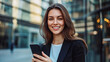 © Michael - Smiling young businesswoman holds her smartphone in a bustling city business district
