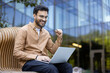 © Liubomir - Young man sitting on bench uses laptop, expressing excitement outdoors. Casual office style, focusing on success and achievement. Contemporary architecture and greenery create work environment.