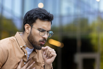 Young man coughing with hand on chest showing signs of discomfort or cold. Image captures concern, health issues in outdoor urban setting. Glasses add to somber mood.