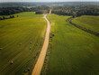© Emvats - Aerial image of a winding dirt road cutting through expansive green fields dotted with hay bales. A dense forest borders the horizon, creating a picturesque rural landscape.