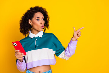 Poster - Photo of astonished woman dressed striped top indicating look at sale empty space hold smartphone isolated on yellow color background