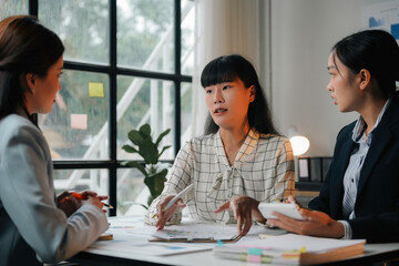 Wall Mural - Three asian businesswomen collaborating at a desk, analyzing financial data, and strategizing for a new business venture