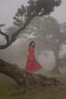 © Milou Dirks - art portrait of asian girl in a long red dress standing on tree trunk in misty landscape of Fanal Forest, Madeira, Portugal
