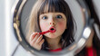 © vita - A little beautiful girl with big gray eyes looks into round mirror carelessly applying red lipstick on her lips. A child with a surprised look on face