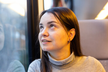 Dark-haired female traveler enjoying journey in modern comfortable train, looking with interest at picturesque scenery outside window