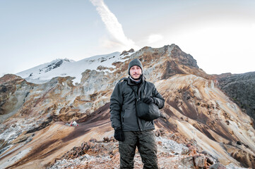 young man smiling in a mountain landscape with a hat for the cold, warm winter clothes in ocher colors and a glacier in the background on a hill